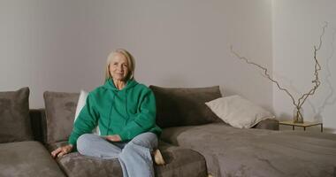 A beautiful elderly woman of European appearance with long gray hair is sitting on a sofa in a modern home interior, her leg bent under her in a shoe and smiling while looking at the camera video