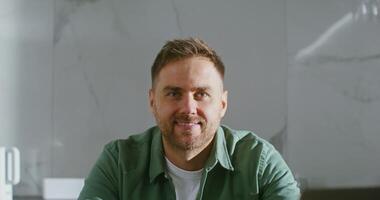 A young stylishly dressed man of European appearance with a short light beard, smiling while looking straight into the camera while sitting at the kitchen table. Portrait video