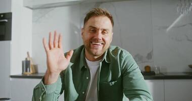 Front camera, a young respectable man of European appearance starts a chat, waving in a sign of greeting, talking to a client when working remotely from home video