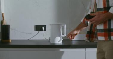 A man includes a white stylish electric kettle, standing on a black marble table top of a modern kitchen and sits at the table waiting. Close-up of a kettle, face men are not visible video
