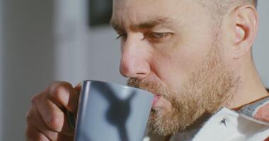 Close-up on the face of a young blond man with a beard drinking a hot drink from a mug and staring at the phone without taking his eyes off. Side videography video
