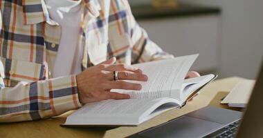 A young man of European appearance, dressed in casual clothes, leafs through a book while sitting in front of an open laptop at the table, close-up male hands, the man's face is not visible video