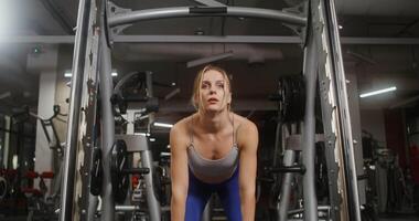 Young beautiful woman with athletic physique, dressed in a sports uniform, doing bends with a barbell on the exercise machine, doing in the gym. Front videography, close-up video