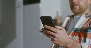 A young bearded man is drinking coffee while sitting in the kitchen and uses a mobile phone. Gradually shifting focus from a man's hand with a phone to a man's face taking a sip from a mug video