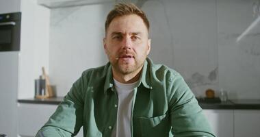 Front camera, a young respectable man communicates with a client via chat, looking directly into the camera, sitting at the table in the kitchen at home video