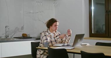 Young curly-haired man of European appearance, dressed in casual clothes in thought, sits in front of open books and a laptop at the table in the kitchen in a modern home interior video