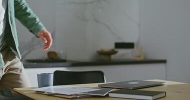 A young man of European appearance sits down at a kitchen table equipped for remote work, and opening the lid of a laptop begins to work with a focused look video