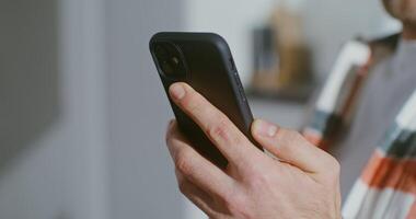 A young bearded man of European appearance is drinking coffee while sitting in the kitchen and holding a mobile phone in his hand. Close-up of a man's hand with a phone, a man's face is blurry video