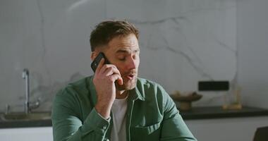 Young respectable man discouraged by a conversation on a mobile phone hangs up in confusion, while sitting at the kitchen table in front of an open laptop video