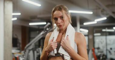 Young beautiful woman of European appearance with a towel around her neck, tired after training, looking directly at the camera while standing in the gym video