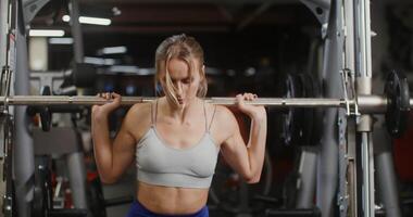 Young beautiful woman of European appearance, dressed in a sports uniform, squats with a barbell on the exercise machine, doing in the gym, close-up video