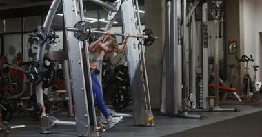 Young beautiful woman of athletic physique, dressed in a sports uniform, squats with a barbell on the exercise machine, doing in the gym. filming from the side, general plan of the gymnasium video