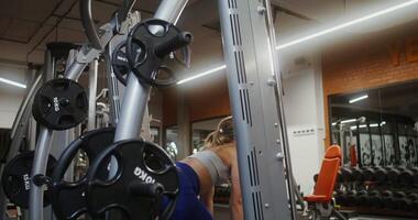 A young woman of blonde athletic physique, dressed in a sports uniform, doing bends with a half-squat with a barbell on an exercise machine for exercises, doing in gym. filming from behind video
