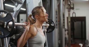 Young beautiful woman, dressed in a sports uniform, squats with a barbell on the exercise machine, doing in the gym. filming from the side, close-up video