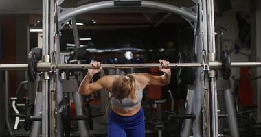 Young blonde woman of european appearance, dressed in a sports uniform, squats with a barbell with a light weight using an exercise machine for exercises, doing in the gym video