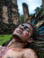 A man with tribal face paint and a feathered headdress rests against a stone structure photo