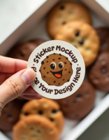 A hand holds a sticker mockup featuring a smiling chocolate chip cookie above a box of assorted cookies psd