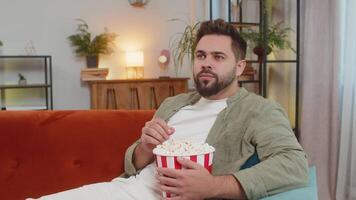 Handsome brunette man eating popcorn and watching movie sitting on sofa couch at home apartment video