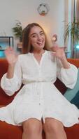 Happy young woman vlogger smiling friendly at camera and waving hands while sitting on couch at home video