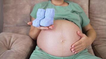 Leaned back pregnant woman on the couch stroking her big belly with one hand. In her other hand she's holding baby booties and shoving them closer to the camera while camera moves back. video