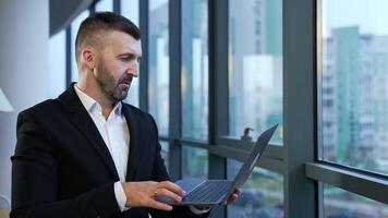 Caucasian man wearing black suit stands in the office and holds the laptop. Man with a personal computer in front of the panoramic window at work. video