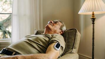 A senior man relaxing in a chair with a smart patch on his upper arm that displays oxygen saturation levels illuminated by soft ambient light from a lamp nearby.. photo