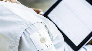 A closeup shot of an advanced chest patch sensor adhered to the skin of an older adult capturing electrical signals with clear visual feedback on a nearby digital tablet.. photo