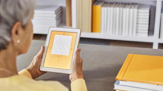 A senior participant surrounded by books and study materials uses a tablet to follow along with an online cognitive exercise a focused expression on their face.. photo