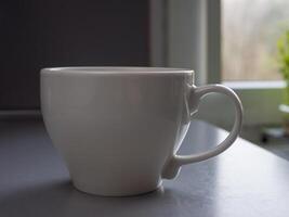 Large white mug brimming with steaming coffee rests on a table beside a window, basking in natural light and creating a warm, inviting atmosphere perfect for morning relaxation photo