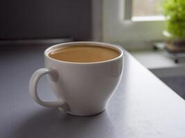 Large white mug full of coffee sitting on a table by a window with a small plant in a pot on the windowsill, enjoying a quiet moment with a warm beverage photo