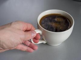 Hand gently grasps the handle of a white mug filled with dark, aromatic coffee, resting on a simple gray table, creating a cozy and inviting scene photo