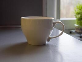 Large white coffee mug resting on a kitchen counter, positioned in front of a window. Out-of-focus green plant on the windowsill adds a touch of nature to the serene, blurred background photo