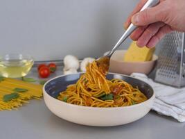 Chef is using a fork to pick up balanesi pasta from a bowl, with ingredients such as raw pasta, mushrooms, tomatoes, cheese, olive oil, basil, and a grater visible in the background photo