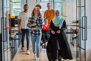 A diverse group of business people walking a corridor in the glass-enclosed office of a modern startup, including a person in a wheelchair and a woman wearing a hijab, showing a dynamic mix of photo