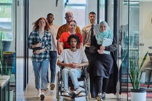 A diverse group of young business people walking a corridor in the glass-enclosed office of a modern startup, including a person in a wheelchair and a woman wearing a hijab, showing a dynamic mix of photo