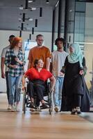 A diverse group of young business people walking a corridor in the glass-enclosed office of a modern startup, including a person in a wheelchair and a woman wearing a hijab, showing a dynamic mix of photo