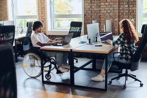 Business colleagues work within a modern startup coworking office, with one of them, an African America in a wheelchair, actively engaging with computers and laptops to collaboratively address various photo