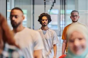 A diverse group of business people walking a corridor in the glass-enclosed office of a modern startup, including a person in a wheelchair and a woman wearing a hijab, showing a dynamic mix of photo