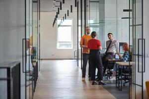 A business group, including an African American businessman in a wheelchair, collaborates within a modern glass office, actively engaged around a computer and laptop, collectively solving diverse photo