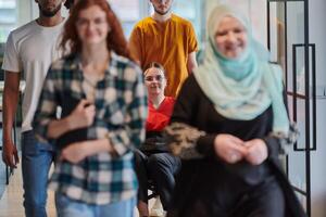 A diverse group of business people walking a corridor in the glass-enclosed office of a modern startup, including a person in a wheelchair and a woman wearing a hijab, showing a dynamic mix of photo