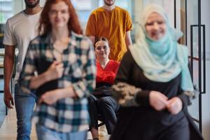 A diverse group of business people walking a corridor in the glass-enclosed office of a modern startup, including a person in a wheelchair and a woman wearing a hijab, showing a dynamic mix of photo