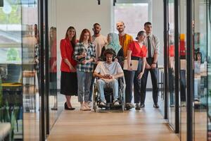 A diverse group of young business people walking a corridor in the glass-enclosed office of a modern startup, including a person in a wheelchair and a woman wearing a hijab, showing a dynamic mix of photo