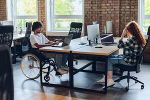 Business colleagues work within a modern startup coworking office, with one of them, an African America in a wheelchair, actively engaging with computers and laptops to collaboratively address various photo