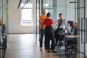 A business group, including an African American businessman in a wheelchair, collaborates within a modern glass office, actively engaged around a computer and laptop, collectively solving diverse photo