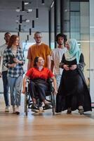 A diverse group of young business people walking a corridor in the glass-enclosed office of a modern startup, including a person in a wheelchair and a woman wearing a hijab, showing a dynamic mix of photo