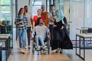 A diverse group of young business people walking a corridor in the glass-enclosed office of a modern startup, including a person in a wheelchair and a woman wearing a hijab, showing a dynamic mix of photo