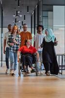A diverse group of young business people walking a corridor in the glass-enclosed office of a modern startup, including a person in a wheelchair and a woman wearing a hijab, showing a dynamic mix of photo