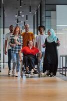 A diverse group of young business people walking a corridor in the glass-enclosed office of a modern startup, including a person in a wheelchair and a woman wearing a hijab, showing a dynamic mix of photo