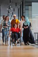 A diverse group of young business people walking a corridor in the glass-enclosed office of a modern startup, including a person in a wheelchair and a woman wearing a hijab, showing a dynamic mix of photo