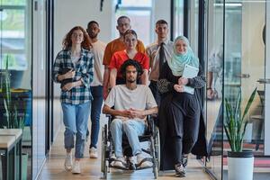A diverse group of young business people walking a corridor in the glass-enclosed office of a modern startup, including a person in a wheelchair and a woman wearing a hijab, showing a dynamic mix of photo